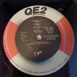 Używane winyle, Płyty winylowe na sprzedaż, sklep winyle online Zbliżenie na Mike Oldfield – QE2 VG- winyl, idealny dla tych, którzy szukają płyt winylowych online. Czarna etykieta z czerwonymi i szarymi akcentami wymienia stronę pierwszą: „Taurus”, „Sheba”, „Conflict” i „Arrival”. Zauważ kultowe logo Virgin na dole. Sklep online z winylami
